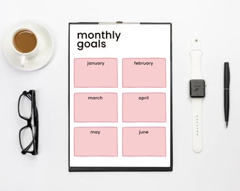 Könnte beinhalten: Ein Klemmbrett mit einem "monatlichen Ziele"-Planer, mit rosa Kästchen für Januar bis Juni. Eine weiße Uhr, ein schwarzer Stift, eine Brille und eine Tasse Kaffee sind um den Planer auf einer weißen Oberfläche angeordnet.