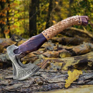 May include: A hand-forged Viking axe with a leather-wrapped handle. The axe head features intricate carvings and runes. The axe is resting on a log in a forest setting.