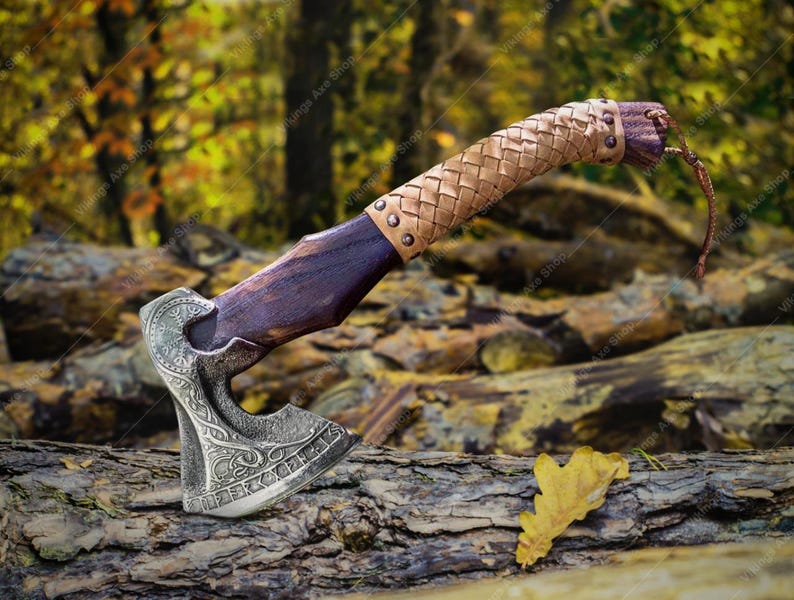 May include: A hand-forged Viking axe with a leather-wrapped handle. The axe head features intricate carvings and runes. The axe is resting on a log in a forest setting.