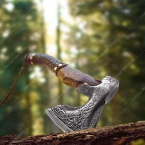 May include: A detailed, silver-colored axe with a wooden handle and leather grip. The axe head features intricate engraved designs. The handle is dark brown with gold-colored rivets. The axe is displayed on a piece of wood against a blurred forest background.