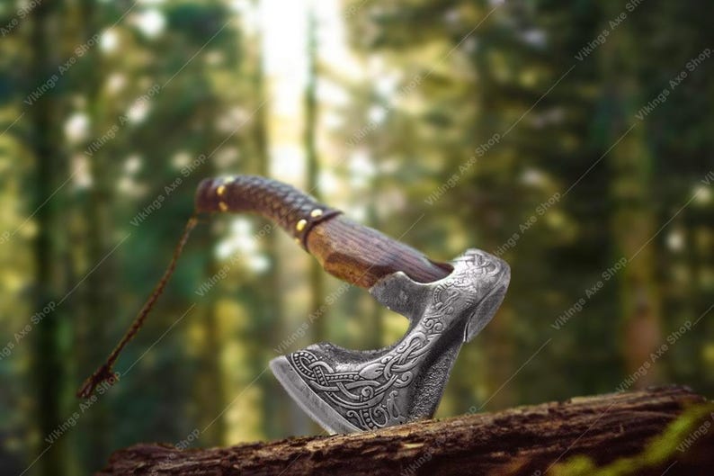 May include: A detailed, silver-colored axe with a wooden handle and leather grip. The axe head features intricate engraved designs. The handle is dark brown with gold-colored rivets. The axe is displayed on a piece of wood against a blurred forest background.