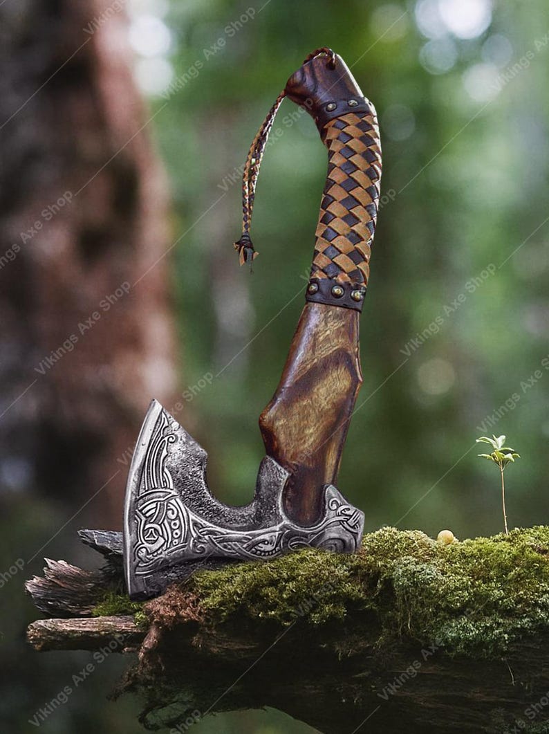 May include: A handcrafted axe with a silver blade featuring intricate Celtic knotwork. The wooden handle is wrapped in a brown and tan woven leather pattern. A leather strap hangs from the top of the handle. The axe is displayed on a moss-covered branch.