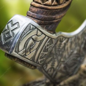 May include: Close-up of a silver axe head with intricate carvings and a wooden handle. The axe head features a geometric pattern and a knotwork design.