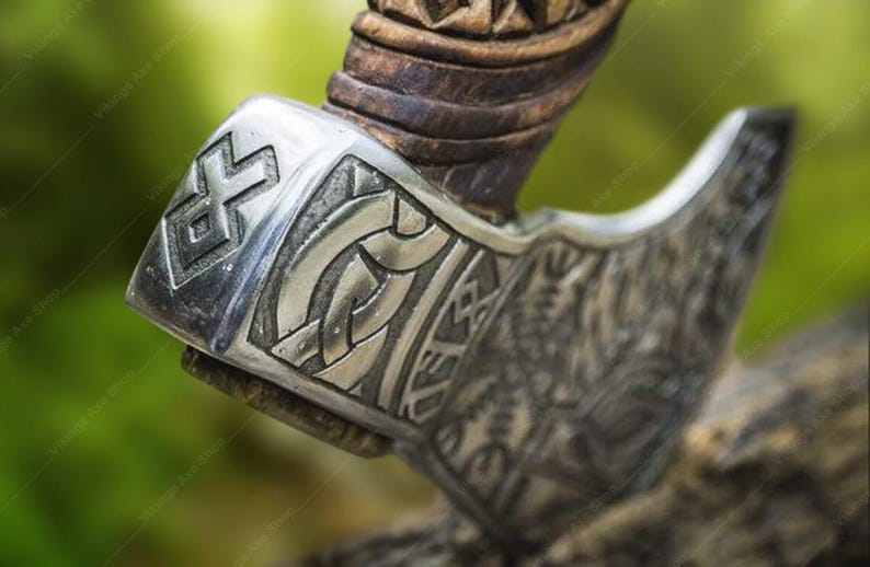May include: Close-up of a silver axe head with intricate carvings and a wooden handle. The axe head features a geometric pattern and a knotwork design.