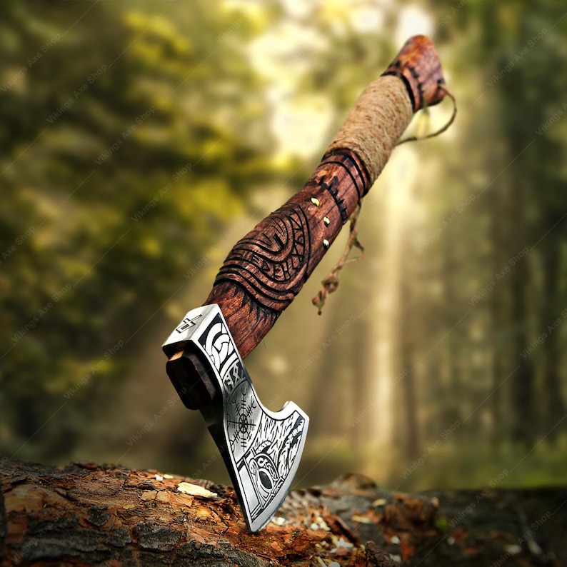 May include: A handcrafted axe with a wooden handle and a metal blade. The handle is wrapped with twine and features intricate carvings. The blade has etched designs. The axe is displayed on a piece of wood against a blurred forest background.