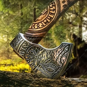 May include: Axe with a wooden handle and a silver-colored head. The handle is carved with intricate patterns, and the axe head features detailed engravings. The axe is displayed on a wooden surface against a blurred forest background.