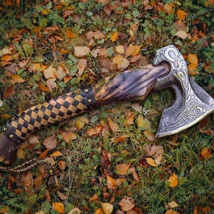 May include: A hand-forged axe with a brown wooden handle and a leather wrap. The axe head has intricate metalwork and a silver finish. The axe is lying on a bed of green grass and fallen autumn leaves.