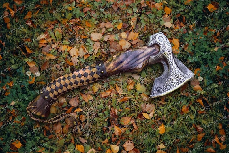 May include: A hand-forged axe with a brown wooden handle and a leather wrap. The axe head has intricate metalwork and a silver finish. The axe is lying on a bed of green grass and fallen autumn leaves.
