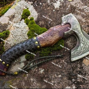 May include: A hand-forged Viking axe with a wooden handle wrapped in leather and a metal head with intricate knotwork designs. The axe is resting on a mossy rock.