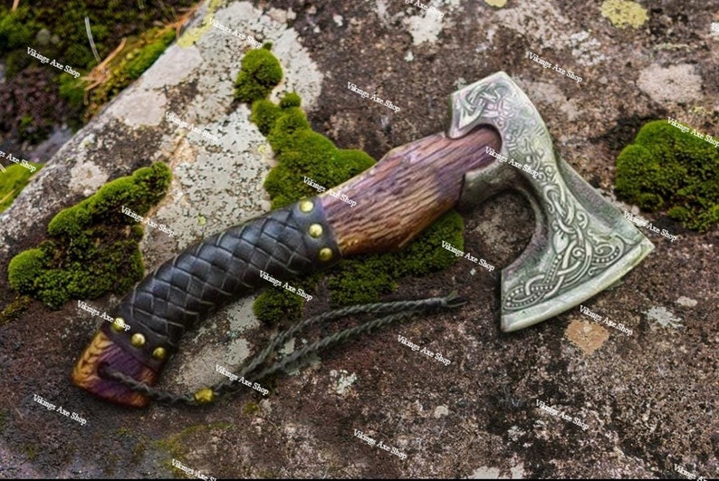 May include: A hand-forged Viking axe with a wooden handle wrapped in leather and a metal head with intricate knotwork designs. The axe is resting on a mossy rock.