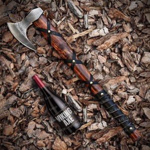 May include: A Viking axe with a wooden handle wrapped in black leather and brass accents. The axe is lying on a bed of wood chips. A wine bottle with a label that reads "Viking Axe" is also in the image.