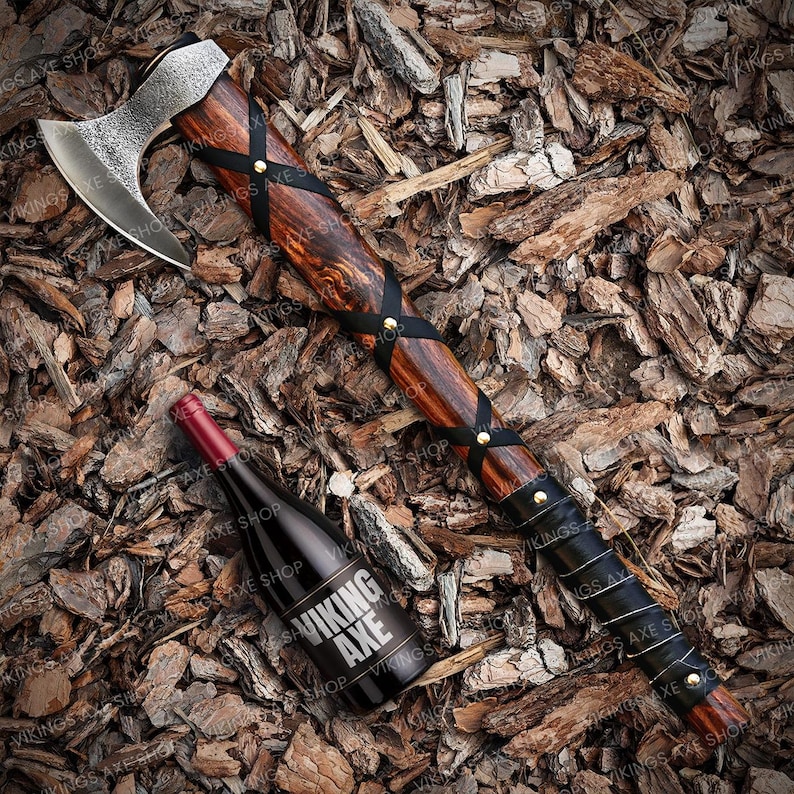 May include: A Viking axe with a wooden handle wrapped in black leather and brass accents. The axe is lying on a bed of wood chips. A wine bottle with a label that reads "Viking Axe" is also in the image.