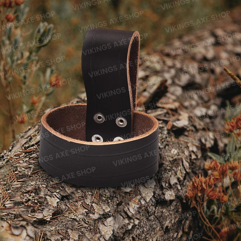 May include: A black leather axe sheath with a brown interior and a loop for attaching to a belt. The sheath has a cylindrical design with a riveted strap and is designed to hold an axe. The text "VIKINGS AXE SHOP" is visible.