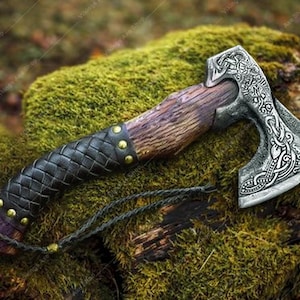 May include: A Viking axe with a wooden handle and a silver-colored head. The handle is wrapped in black leather with gold-colored studs. The axe head features intricate engraved designs. A braided cord is attached to the handle.