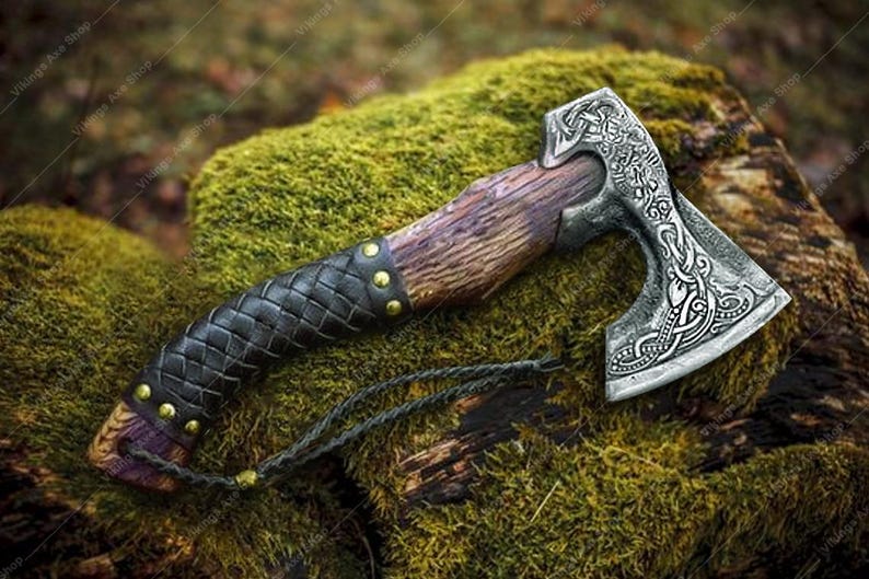 May include: A Viking axe with a wooden handle and a silver-colored head. The handle is wrapped in black leather with gold-colored studs. The axe head features intricate engraved designs. A braided cord is attached to the handle.