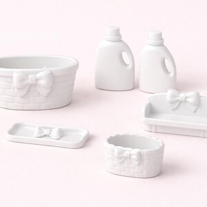 May include: A collection of white ceramic decorative pieces, including a basket, two detergent bottle-shaped items, a small sofa, a tray, and a small oval basket. Each piece features a bow accent, creating a cohesive and charming aesthetic.