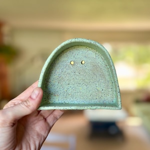 May include: A semi-circular ceramic dish in a light mint green color with a speckled texture throughout. The dish features two small, circular gold-toned studs positioned near the center of the interior surface, designed for holding jewelry.