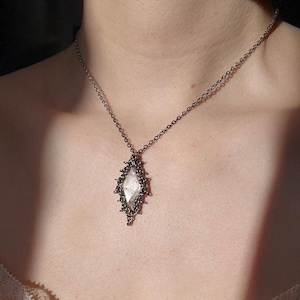 Puede incluir: Un collar con una cadena fina de metal oscuro y un colgante central. El colgante tiene una piedra transparente en forma de rombo, engastada en un marco de metal oscuro y texturizado con pequeñas protuberancias en forma de espinas.