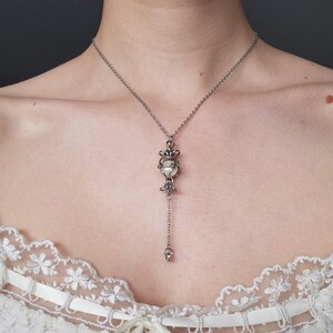 Puede incluir: Un collar de cadena en tono plateado con un colgante central compuesto por anillos de metal entrelazados que encierran una cuenta esférica blanca. El diseño del colgante incluye elementos metálicos decorativos encima y debajo de la cuenta, con un segmento de cadena corto que desciende desde la base y termina en un pequeño anillo de metal.