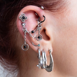 Peut inclure: Une chaîne de cartilage d'oreille argentée avec un clou en forme de trèfle à quatre feuilles orné d'une pierre noire, reliée à un anneau avec une perle noire. L'oreille présente plusieurs piercings dont une petite barre courbée, un anneau avec une croix pendante, une bague et un anneau en croissant texturé.
