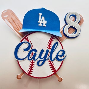 May include: A baseball-themed cake topper with a blue and white LA Dodgers baseball cap, a baseball with the name "Cayle" written on it, a silver number 8, and two crossed wooden baseball bats.