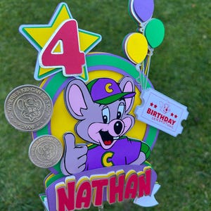 Op de afbeelding: Kleurrijke taarttopper met een cartoon muis karakter in paarse en groene kleding. De topper toont het nummer 4, ballonnen en de naam "Nathan". Er zijn ook twee munten met de tekst "Where a kid can be a kid".
