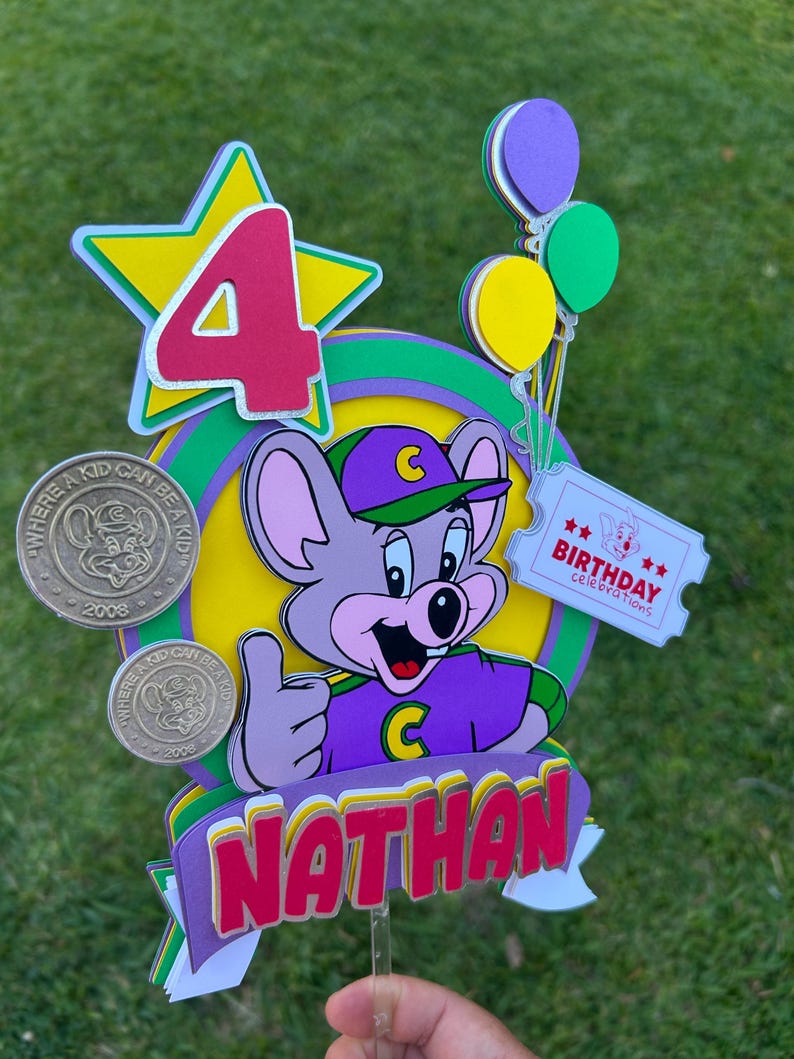 Op de afbeelding: Kleurrijke taarttopper met een cartoon muis karakter in paarse en groene kleding. De topper toont het nummer 4, ballonnen en de naam "Nathan". Er zijn ook twee munten met de tekst "Where a kid can be a kid".