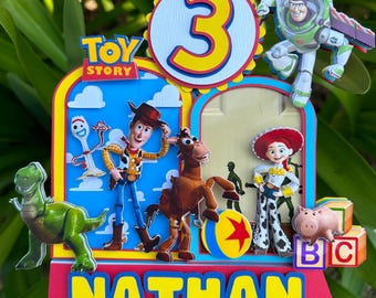 Könnte beinhalten: Ein farbenfroher "Toy Story"-Kuchenaufsatz mit der Zahl 3, dem Namen "Nathan" und Charakter-Ausschnitten von Woody, Buzz Lightyear, Jessie, Rex und anderen. Der Aufsatz ist vor einem Hintergrund aus grünem Laub angebracht.