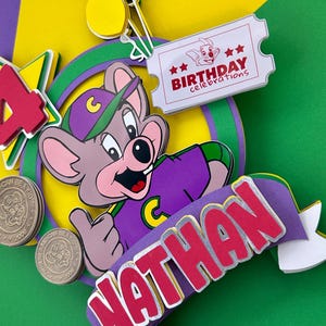 Op de afbeelding: Kleurrijke taarttopper met een cartoonmuis in een paars shirt en pet. De topper bevat de naam "NATHAN", een ticket met "BIRTHDAY celebrations" en het cijfer "4".