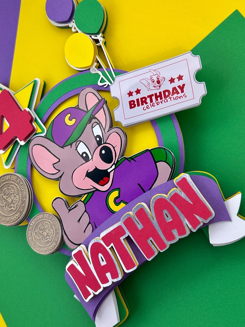 Op de afbeelding: Kleurrijke taarttopper met een cartoonmuis in een paars shirt en pet. De topper bevat de naam "NATHAN", een ticket met "BIRTHDAY celebrations" en het cijfer "4".
