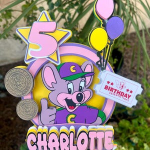 Op de afbeelding: Kleurrijke taarttopper met een cartoon muis, het nummer 5 in een ster, ballonnen en de naam "Charlotte". Inclusief een ticket met de tekst "Birthday Celebrations" en twee munten.