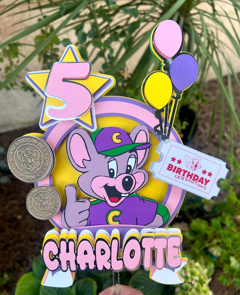 Op de afbeelding: Kleurrijke taarttopper met een cartoon muis, het nummer 5 in een ster, ballonnen en de naam "Charlotte". Inclusief een ticket met de tekst "Birthday Celebrations" en twee munten.