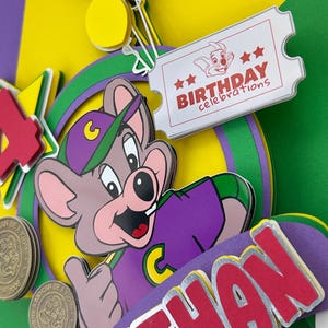 Op de afbeelding: Een kleurrijke verjaardagskaart met een cartoon muis karakter in een paars shirt en groene pet. De kaart bevat de naam "Nathan" in rode letters, een ticket met "Birthday Celebrations" en een rode "4".