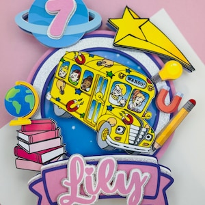 May include: A colorful, layered cake topper featuring a yellow school bus with students, a pink number 7, a shooting star, and the name "Lily". Additional elements include a globe, books, and a pencil.