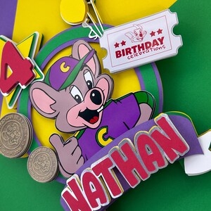 Op de afbeelding: Een kleurrijke taarttopper voor een verjaardag met een stripfiguur in een paars shirt en hoed. De topper bevat de naam "Nathan" in rode letters, een ticket met "Birthday celebrations" en het nummer "4". Er zijn ook twee munten.