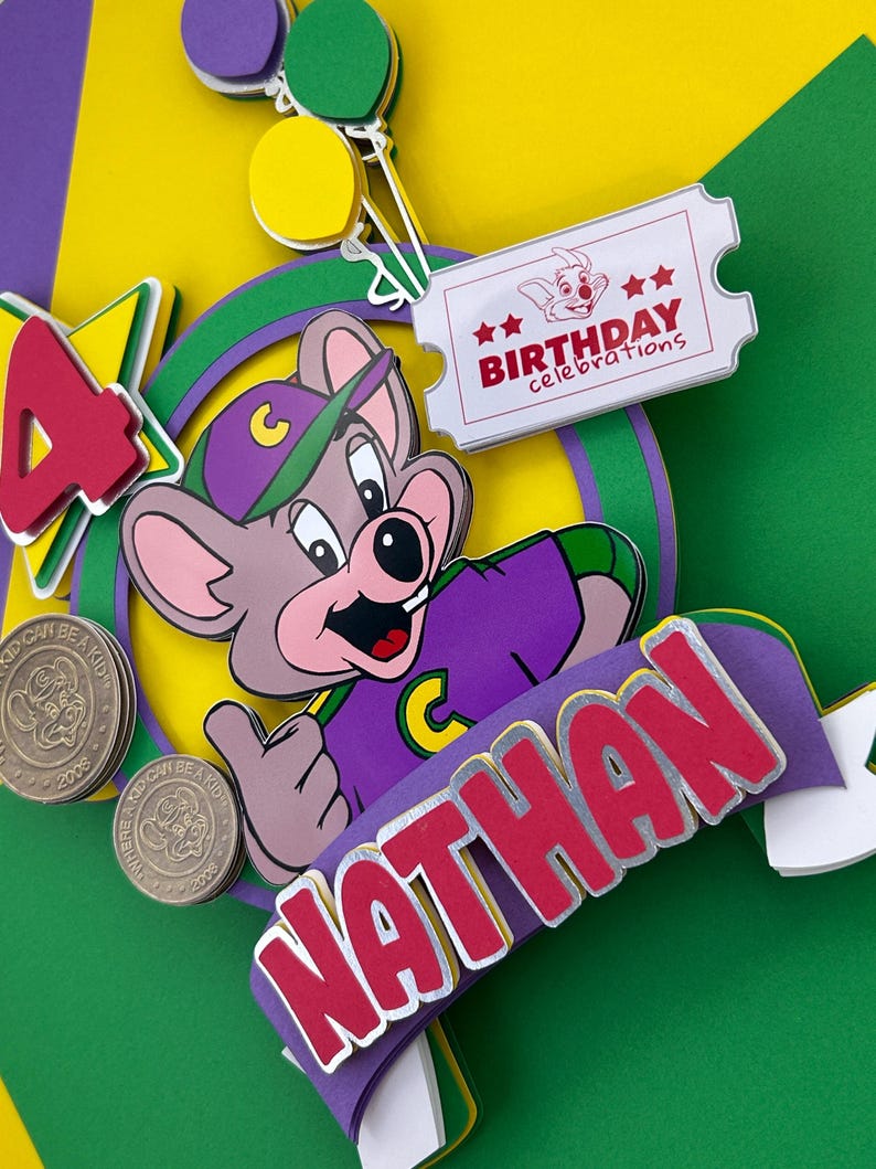 Op de afbeelding: Een kleurrijke taarttopper voor een verjaardag met een stripfiguur in een paars shirt en hoed. De topper bevat de naam "Nathan" in rode letters, een ticket met "Birthday celebrations" en het nummer "4". Er zijn ook twee munten.
