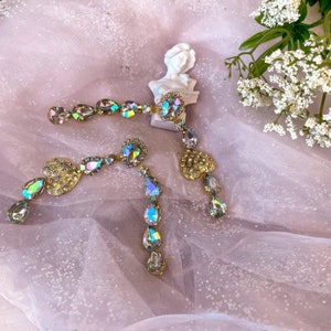 Peut inclure: Une paire de boucles d'oreilles pendantes en cristal iridescent avec des breloques en forme de cœur. Les boucles d'oreilles sont dorées et présentent des cristaux en forme de goutte.