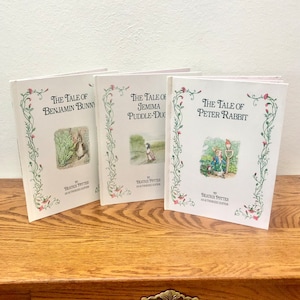Peut inclure: Trois livres reliés vintage avec des couvertures blanches et des bordures florales vertes. Les titres sont "The Tale of Benjamin Bunny", "The Tale of Jemima Puddle-Duck" et "The Tale of Peter Rabbit". Chaque livre présente une illustration du personnage principal sur la couverture.