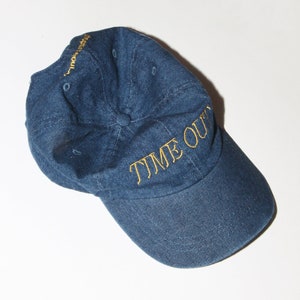 May include: A dark blue denim baseball cap with a gold embroidered inscription that reads "TIME OUT".
