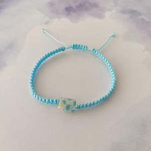 Puede incluir: Una pulsera de macramé azul claro con una cuenta cuadrada transparente. La cuenta presenta un diseño floral con flores amarillas y azules. La pulsera es ajustable.