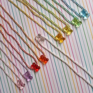 Puede incluir: Un conjunto de 10 collares de ositos de gominola de colores. Cada collar está hecho con pequeñas cuentas blancas y tiene un dije de osito de gominola de un color diferente. Los collares están dispuestos en forma de arcoíris.
