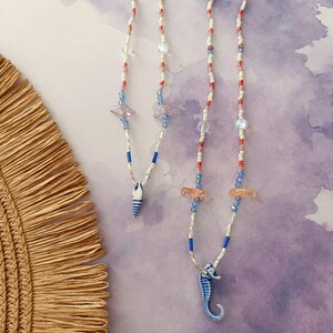Puede incluir: Dos collares de cuentas con colgantes de caballitos de mar y conchas. Los collares presentan cuentas blancas, rojas y azules, con colgantes de caballitos de mar azules y blancos y abalorios de conchas. El fondo es de color lila claro.