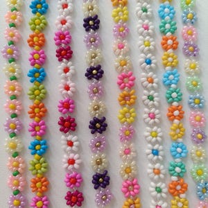 Puede incluir: Una colección de pulseras de cuentas coloridas con diseños de flores de margarita. Las pulseras están hechas con cuentas pequeñas y redondas en varios colores, incluyendo rosa, azul, verde, morado y blanco.