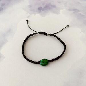 Puede incluir: Una pulsera de cordón negro que presenta una cuenta ovalada de color verde y negro jaspeado en el centro. La pulsera está construida con un diseño de cordón trenzado e incluye un cierre de nudo deslizante ajustable con dos extremos.