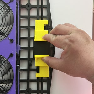 May include: A black plastic frame with two yellow plastic pieces attached to it. The frame has a grid pattern and a black rod running through it. The yellow pieces are rectangular and have a small hole in the center.