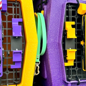May include: Two pet carriers, one yellow and one purple, are stacked on top of each other in the back of a vehicle. The yellow carrier has a purple latch and a green leash attached to it. The purple carrier has a yellow latch.