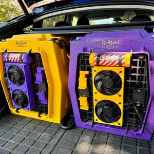 Pu&ograve; includere: Due trasportini per animali domestici con ventilatori incorporati. Il trasportino a sinistra &egrave; giallo con dettagli viola e il trasportino a destra &egrave; viola con dettagli gialli. Entrambi i trasportini hanno la scritta "RuffLand Performance Kennels" stampata su di essi.