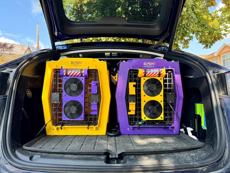 Two Ruffland Performance Kennels in the trunk of a car, each equipped with dual dog crate fans mounted on the door. The left crate is yellow with a purple fan housing, and the right crate is purple with a yellow fan housing. Both feature bolt-on installation, enclosed high-speed fans, and K9 emergency info canisters. Ideal cooling setup for safe dog transport, providing airflow and ventilation during travel or crate use.