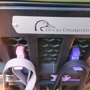 Puede incluir: Un estante de metal negro con dos clips morados y rosados que sujetan dos correas. El estante tiene una placa plateada con un logotipo de pato y el texto "Ducks Unlimited".