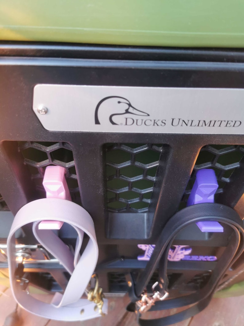 Puede incluir: Un estante de metal negro con dos clips morados y rosados que sujetan dos correas. El estante tiene una placa plateada con un logotipo de pato y el texto "Ducks Unlimited".